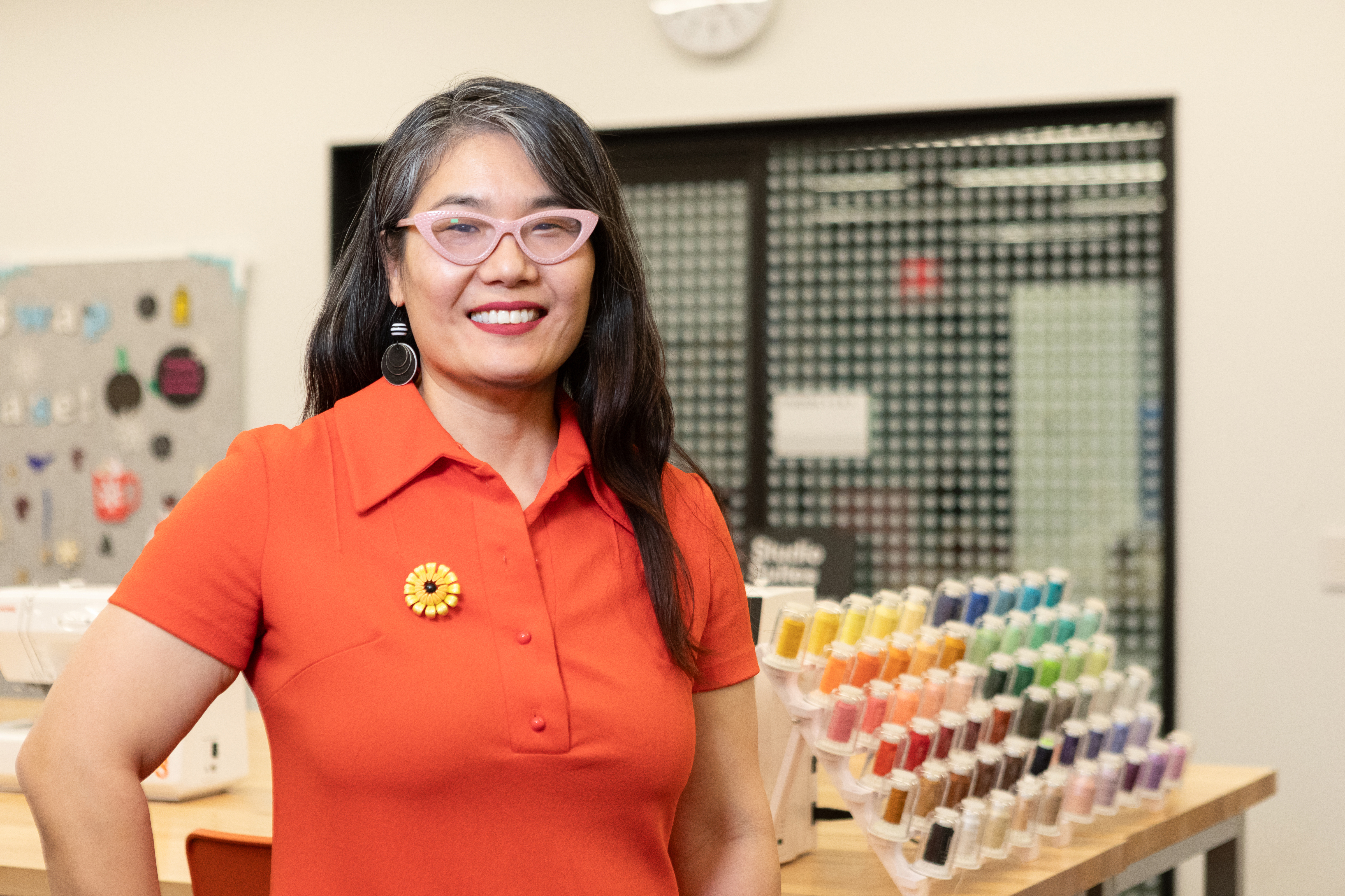 Julie Oh smiling and standing in front of a display of colorful sewing bobbins Julie Oh smiling and standing in front of a display of colorful sewing bobbins