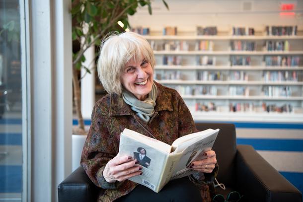 An older woman sits in a chair holding a book called "The Goldfinch" and smiles at the camera. She is wearing a brown patterned jacket, a green scarf, and has a shelf of books behind her and a window to her right.