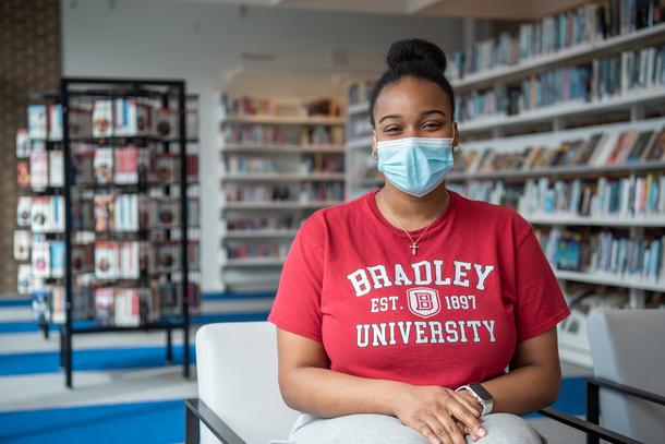 A young woman wearing a mask, a Bradley University red t-shirt, and her hair in a bun sits in a chair with colorful shelves of books behind her.