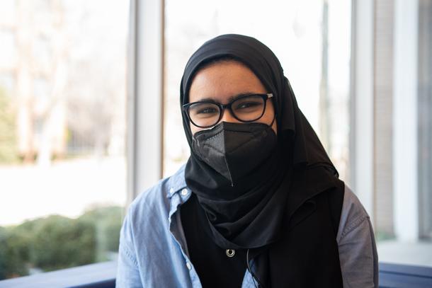 A teenager with glasses, a mask, and a black hijab sits in front of a bright window.