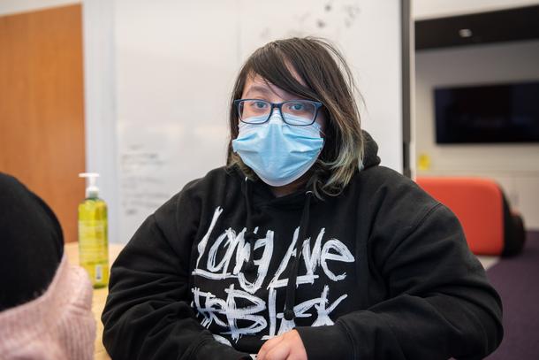 A teenager with glasses, brown hair with blond streaks, and a mask is sitting at a table and wearing a black hoodie with white text on the front.