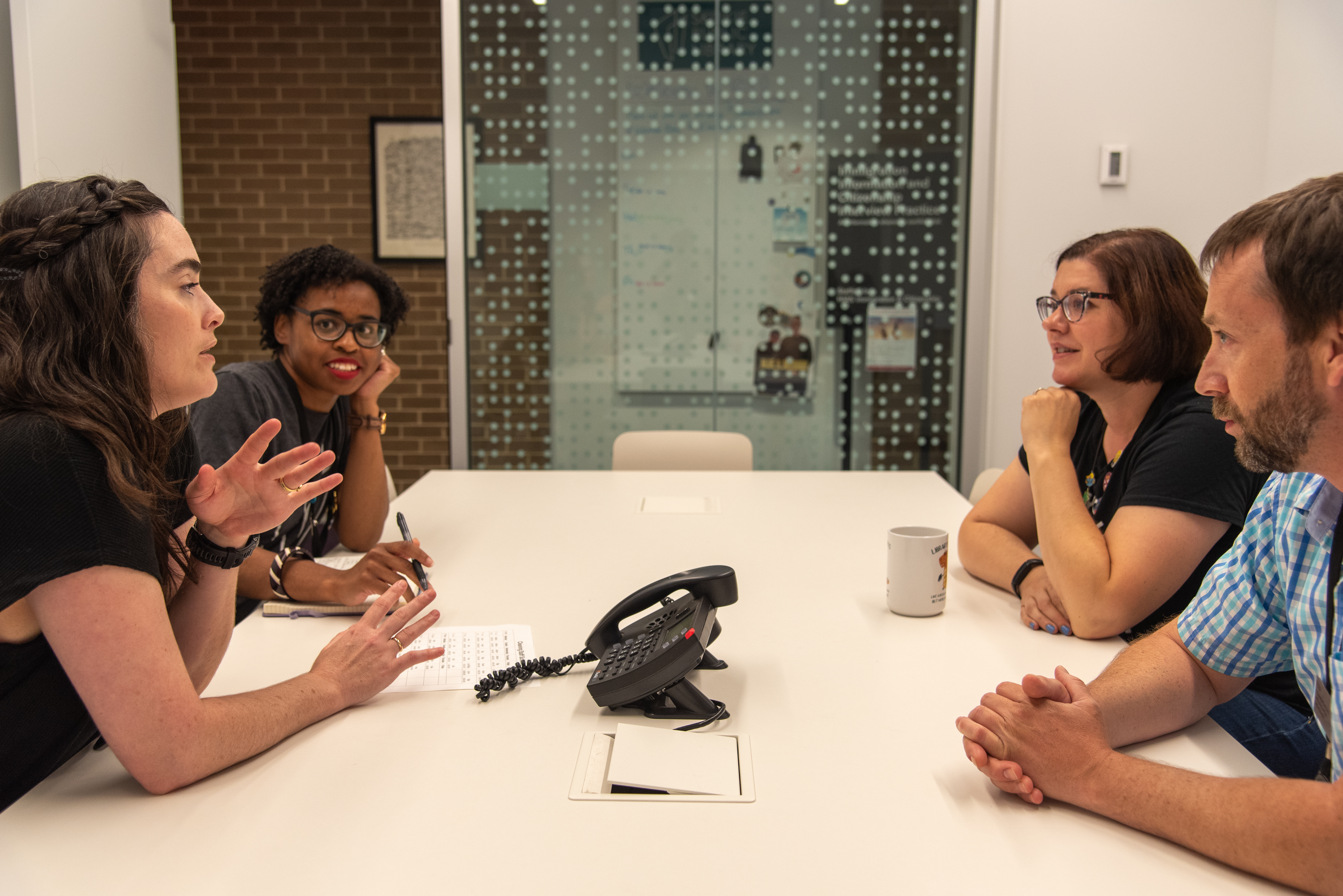 Amber, Katie, Caitlin, and Paul are seated around a table talking. Katie is gesturing. 