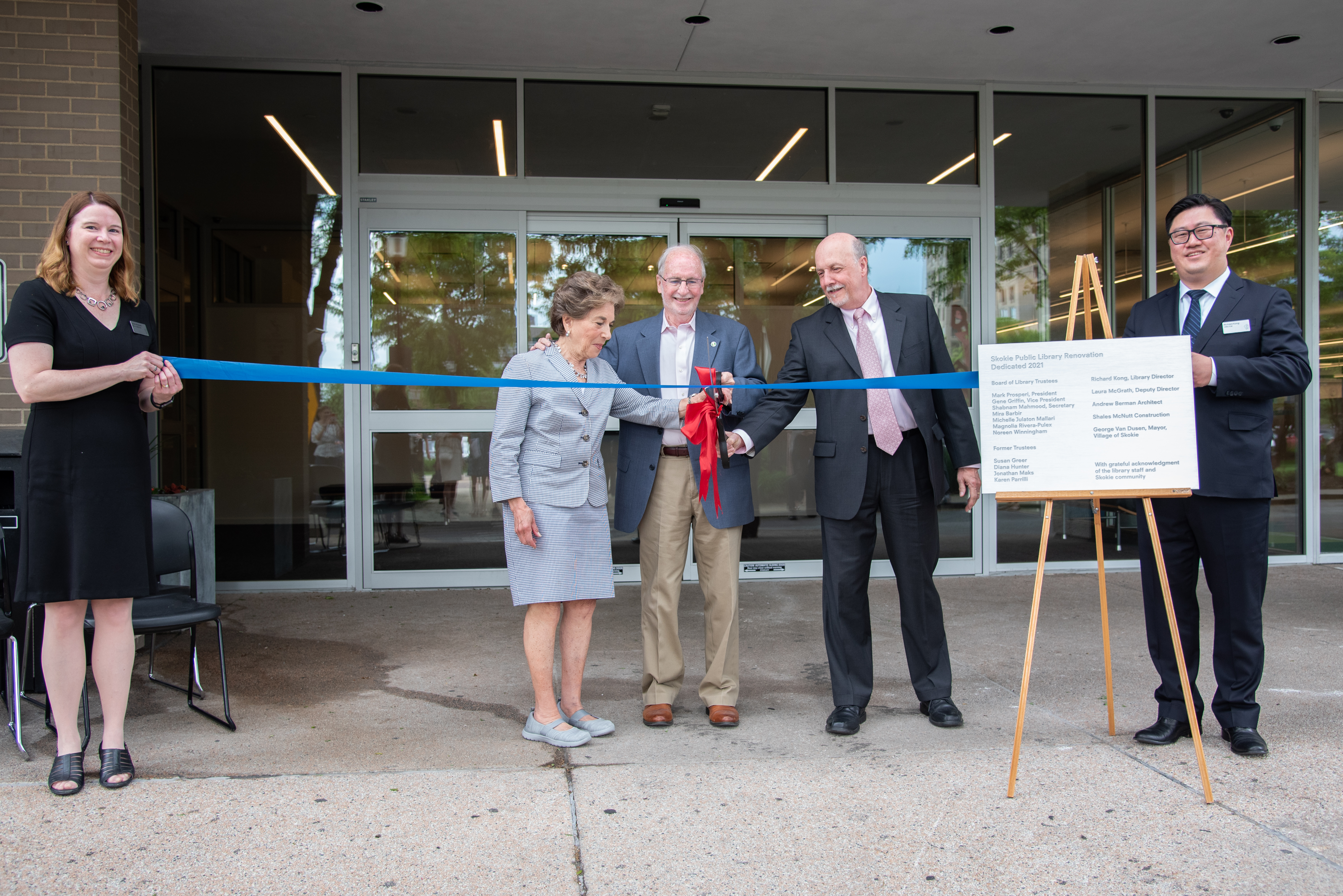 A group of three people wearing business suits use an oversized pair of scissors to cut a ribbon being held by two others A group of three people wearing business suits use an oversized pair of scissors to cut a ribbon being held by two others
