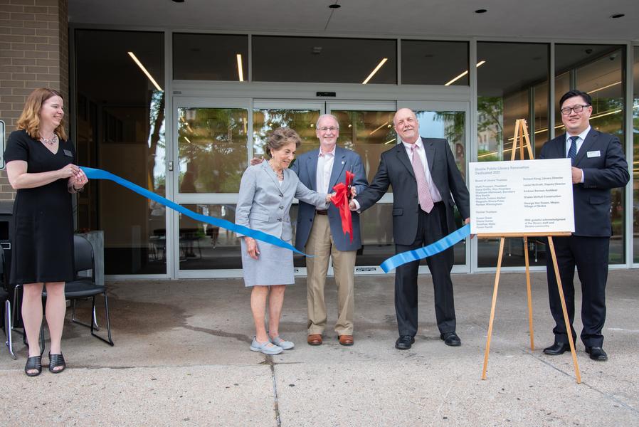 A photo of a ceremonial ribbon cutting. Library Director Richard Kong and Deputy Director Laura McGrath hold a blue ribbon between them, which is falling down after being cut with oversized ceremonial scissors held jointly by library trustee Gene Griffin, Mayor Van Dusen, and Congresswoman Schakowsky.