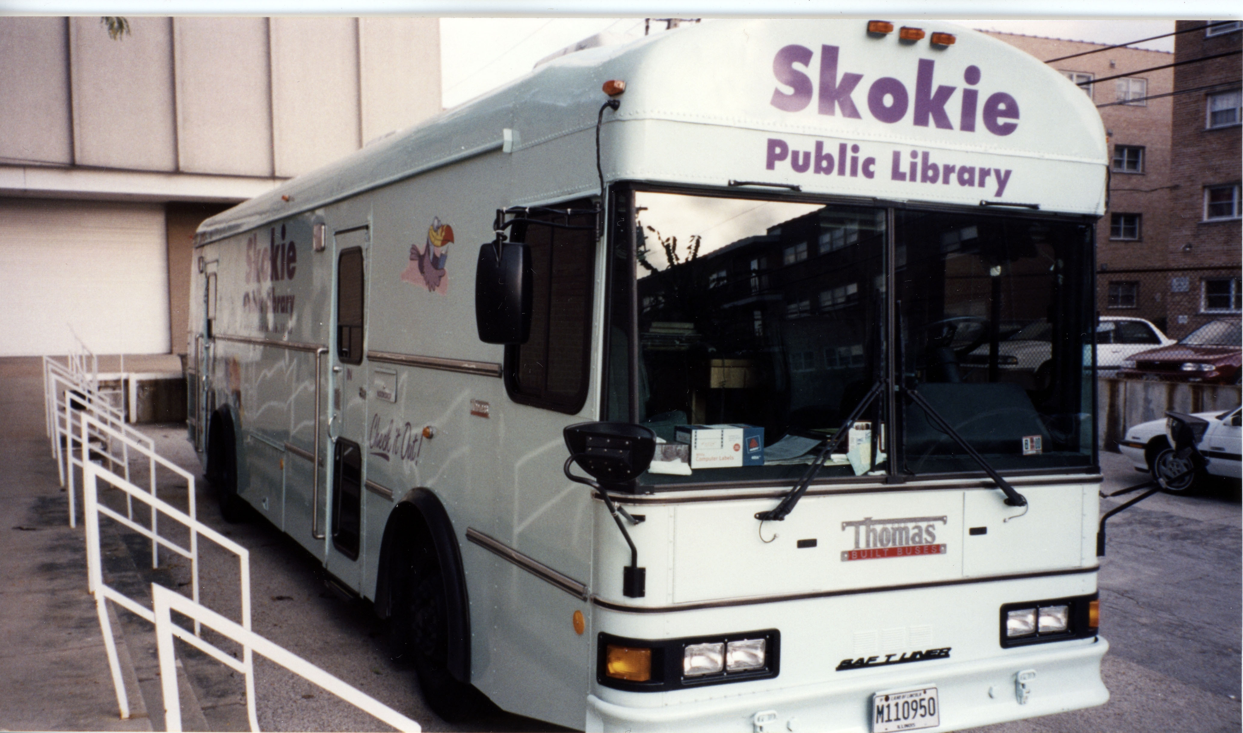 The fourth version of the Skokie Public Library bookmobile, which is white with purple lettering above the windshield, reading "Skokie Public Library".