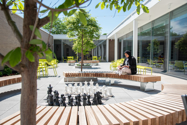 A woman dressed in black sits reading a book on a curved wooden bench in the South courtyard. Around her there is green furniture, green trees, and a giant chess game.