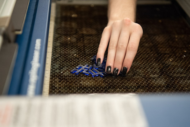 A hand with black nail polish reaches onto the brown grate of the Studio's laser engraver, removing blue angular pieces of acrylic that make up a puzzle.