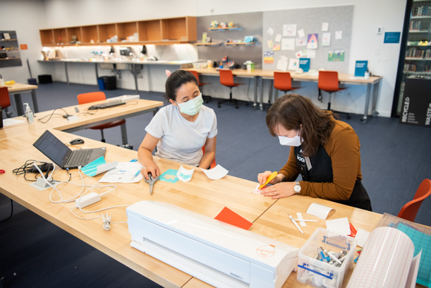 Two masked people, a library patron and a Skokie Public Library staff member, sit at a table in the Studio makerspace of the Skokie Public Library. The staff member is teaching the patron how to use and work with the crafting materials and equipment on the table.