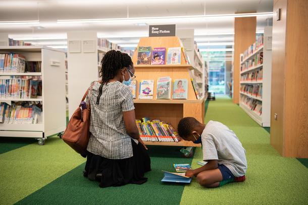 A woman sits in front of a display shelf of kids books while her son reads a book on the green striped carpet.