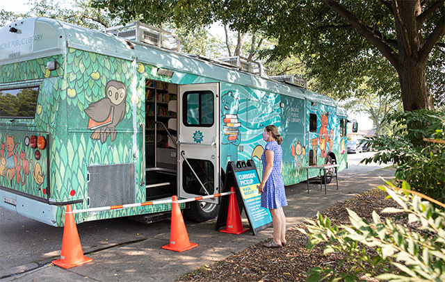 A masked person in a blue dress stands outside the open back door of the Skokie Public Library bookmobile.