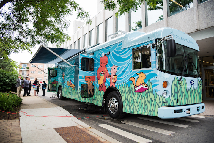 The current Skokie Public Library bookmobile, parked outside of the east entrance to the library. The bookmobile is blue with green grass and yellow and orange animals painted on the side.