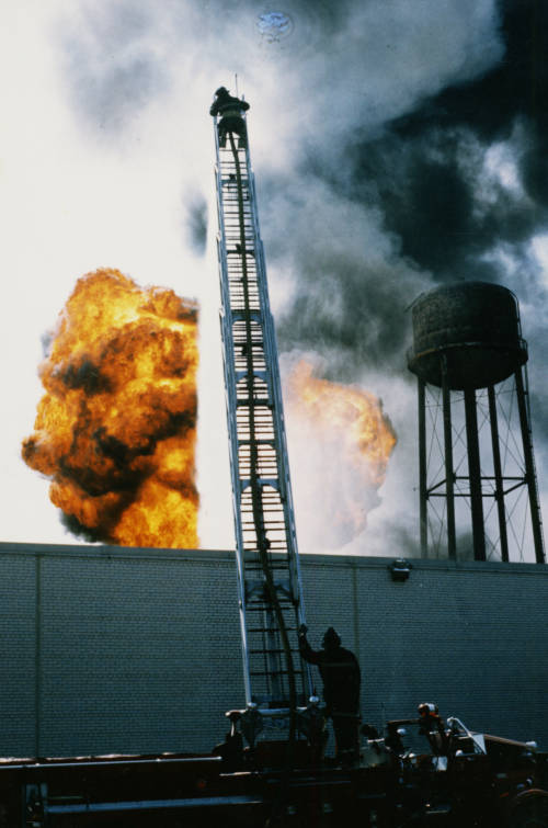 firefighter on top of a tall ladder above a large fire. 