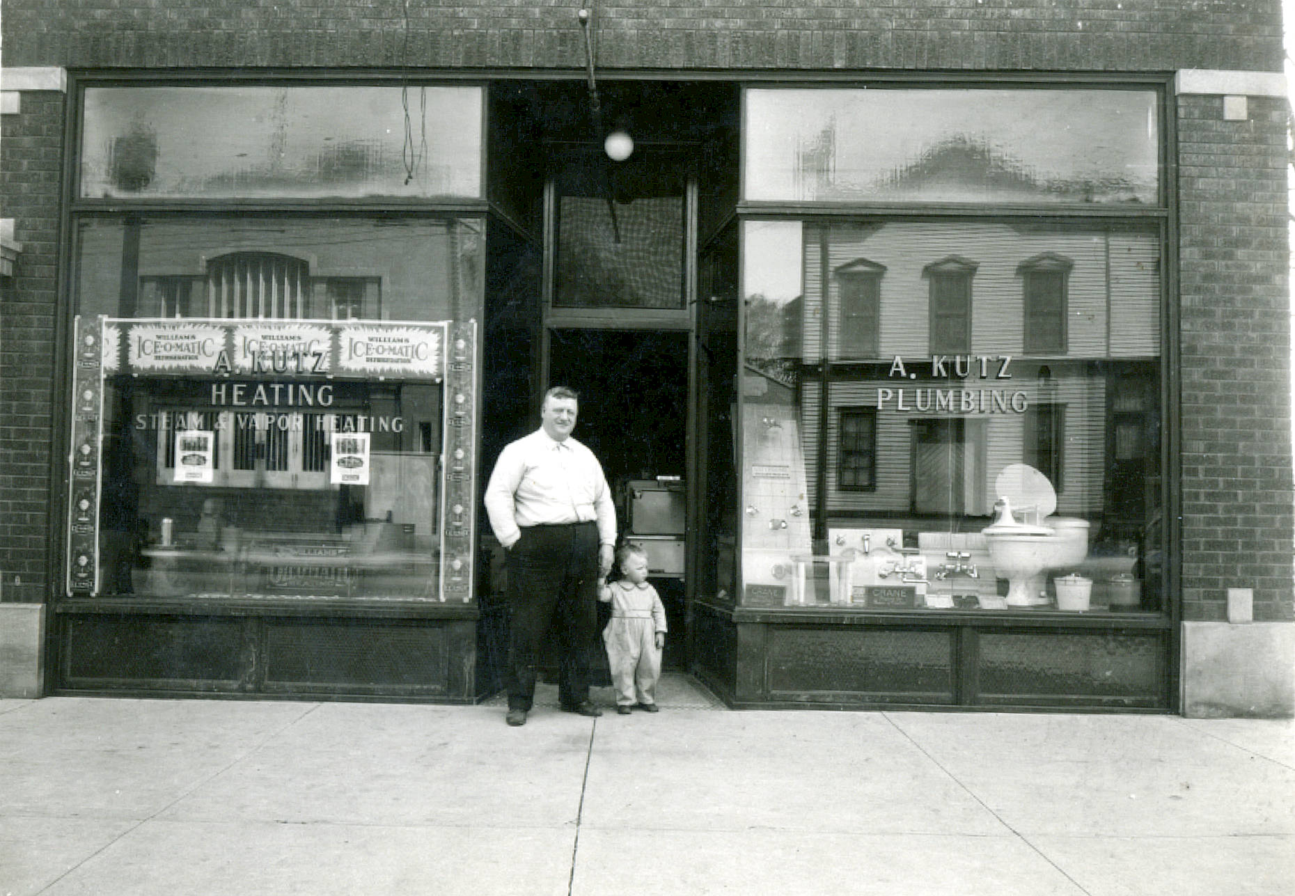 	 Black-and-white photograph of the A. Kutz Plumbing and Heating storefront in Niles Center, Illinois in 1910. An adult man and a toddler are standing in front of the building. 